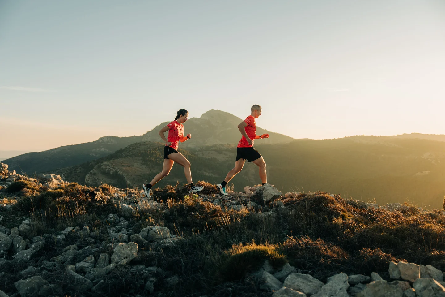 Dos corredores con camisetas rojas recorriendo una cresta de montaña con luz de atardecer