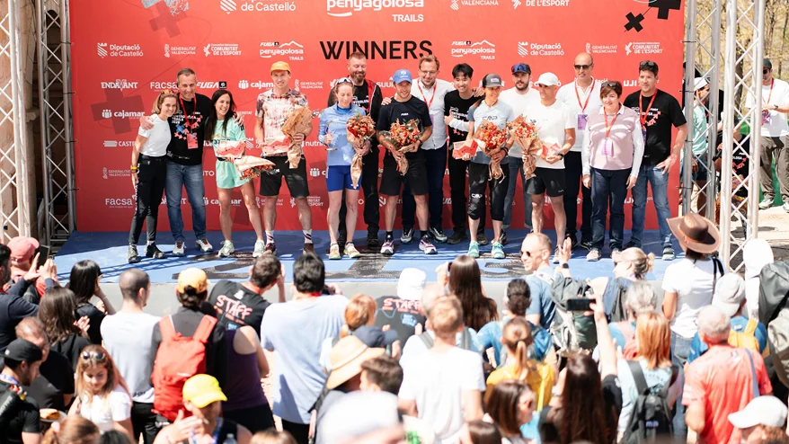 Grupo de participantes con flores y medallas en un podio ante un fondo rojo, con público en primer plano
